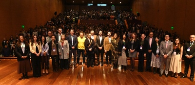Grupo de profesionales posando en un auditorio con escenario de madera, frente a un público numeroso. Visten trajes formales y credenciales, en lanzamiento de AVANZATEC 2026 con iluminación cálida.