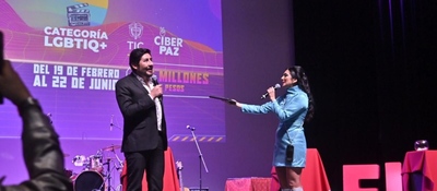 En un escenario iluminado, una presentadora interactua con el vicemninistro frente a una pantalla que anuncia la categoría LGBTIQ+ y fechas del evento. Al fondo se ven instrumentos y mesas rojas.