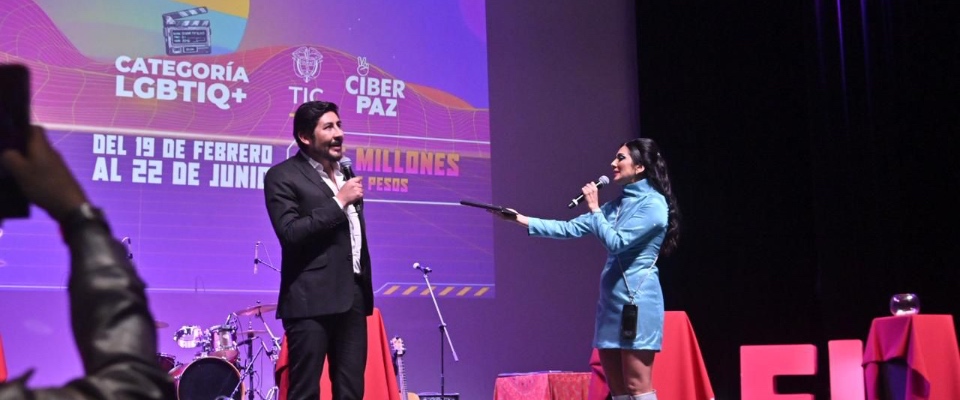 En un escenario, una presentadora conversa con el viceministro TIC frente a una pantalla del evento que anuncia la categoría LGBTIQ+. Al fondo hay instrumentos y mesas rojas.