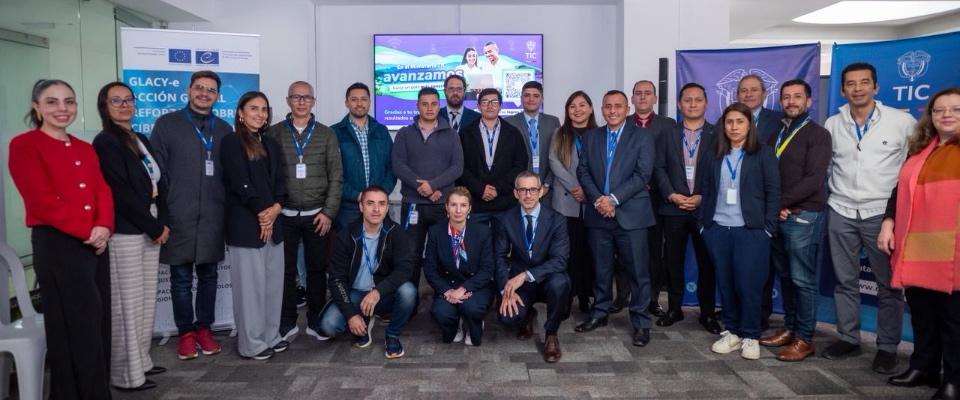 Grupo de profesionales en foto oficial durante actividad institucional del Ministerio TIC, con banners de cooperación y temática de ciberseguridad