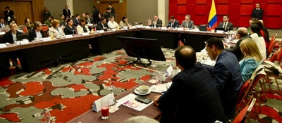 Funcionarios y asistentes participan en reunión oficial alrededor de mesa en U, con bandera de Colombia y pantallas al centro