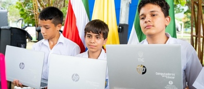 Tres estudiantes con uniforme sostienen portátiles del programa Computadores para Educar frente a banderas, durante entrega de equipos TIC