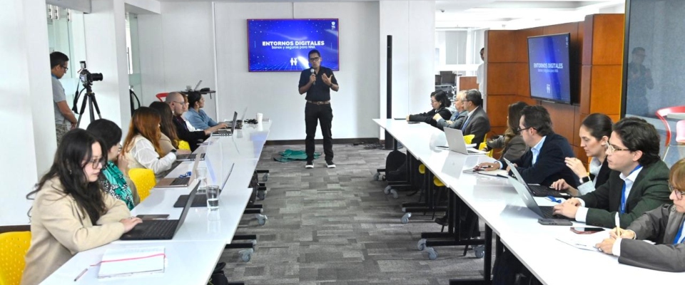 Sala de reuniones: un expositor presenta “Entornos Digitales” frente a una pantalla mientras funcionarios escuchan y trabajan con portátiles en mesas largas.