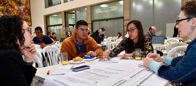 Cuatro personas dialogan en una mesa con documentos y laptops durante un taller; detrás, más grupos trabajan en construir la política de educación digital.