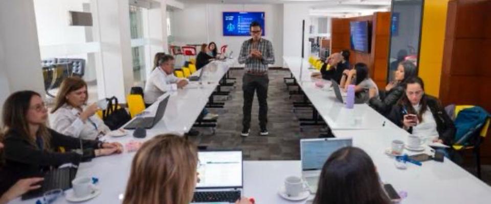 Personas trabajan con portátiles alrededor de una mesa larga en una sala moderna; un hombre de pie habla mientras el grupo participa en la discusión