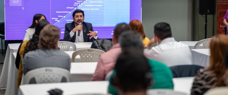 Hombre con micrófono expone ante público sentado; detrás, pantalla con gráficos sobre innovación y transformación digital en evento Hombre con micrófono expone ante público sentado; detrás, pantalla con gráficos sobre innovación y transformación digital en evento