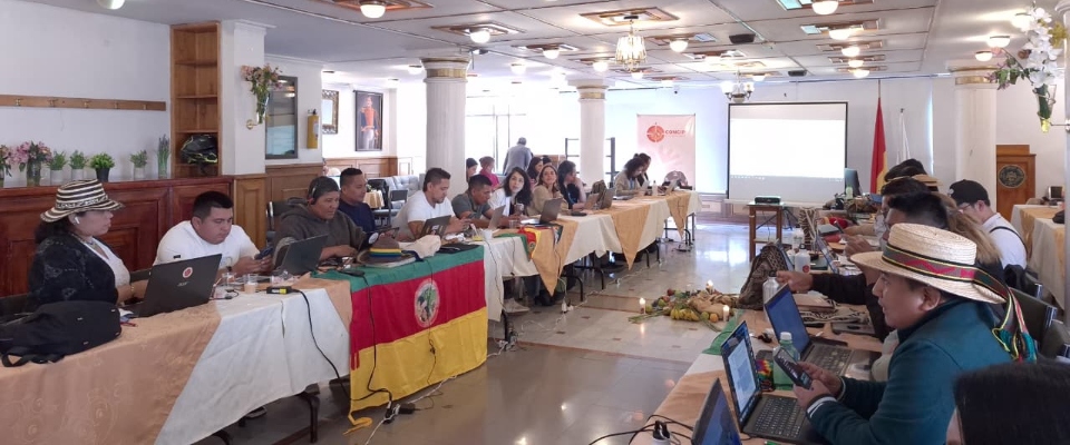 Personas indígenas y funcionarios trabajan con portátiles en mesa larga, en reunión sobre derechos comunicativos liderada por MinTIC
