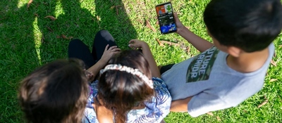 Tres niños sentados en el césped miran un celular. Disfrutan juntos al aire libre mientras exploran contenido digital.