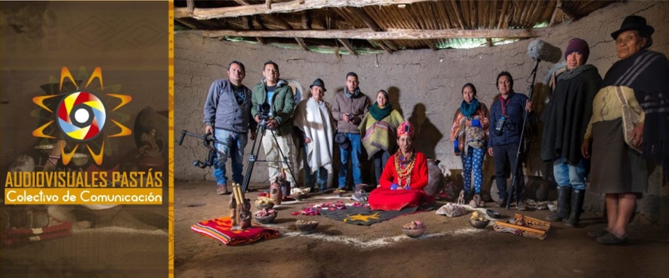 Grupo diverso en vivienda rural indígena; personas con cámaras rodean a mujer en traje tradicional y elementos rituales en el suelo.