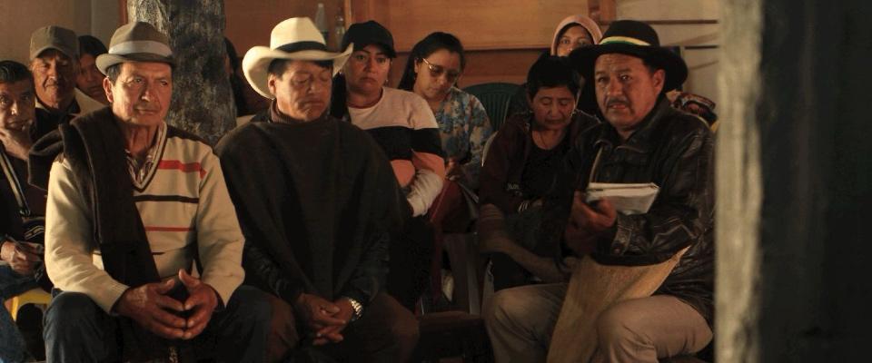 Grupo de hombres y mujeres indígenas sentados en reunión comunitaria; uno habla con cuaderno en mano mientras otros escuchan atentos.