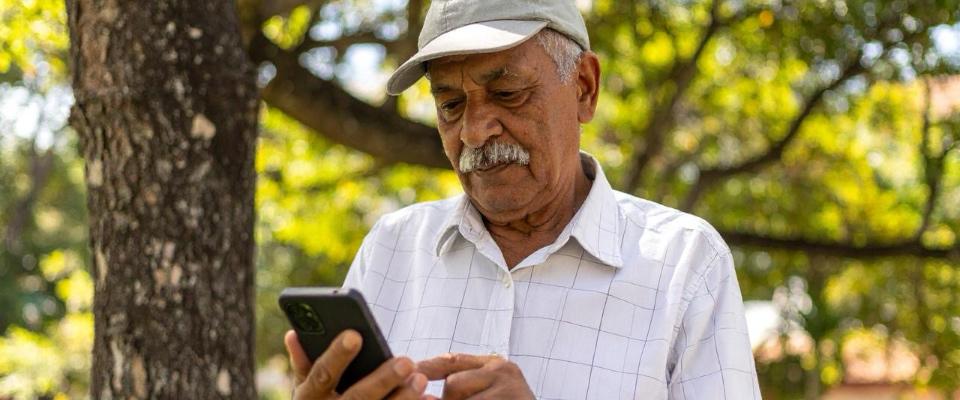 Hombre mayor usa celular bajo un árbol. Representa acceso a telecomunicaciones y oportunidades para servicios comunitarios en banda 900 MHz.