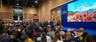 Auditorio lleno en evento oficial; en tarima, pantalla muestra ciudad y lema “Colombia, el país de la belleza” ante asistentes atentos.
