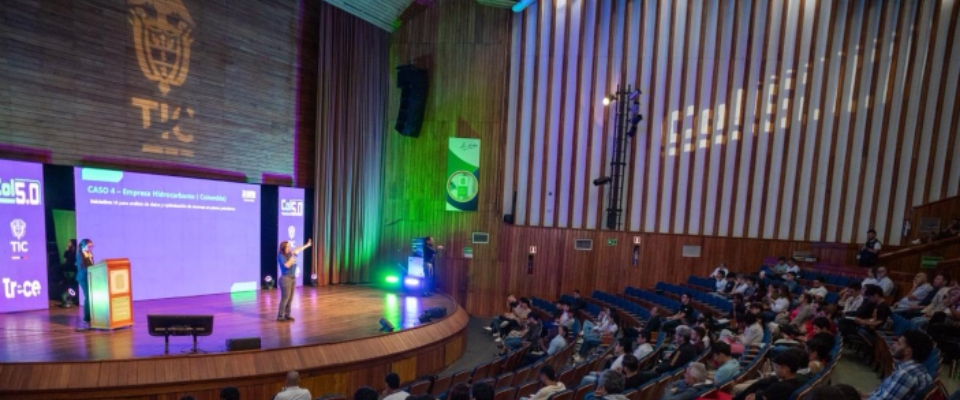 Auditorio amplio con escenario iluminado; ponente expone sobre Colombia 5.0 ante público sentado, con logos TIC y pantallas digitales.