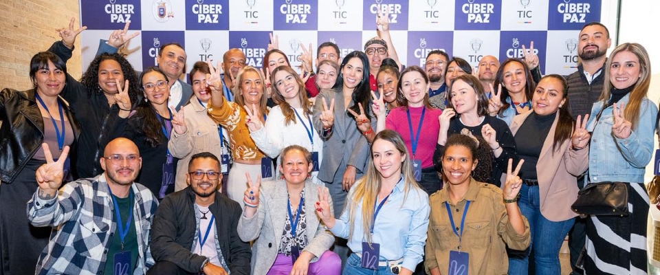 Grupo diverso de personas sonríe frente a fondo “CiberPaz TIC”, levantando signos de paz; ambiente colaborativo por la inclusión digital