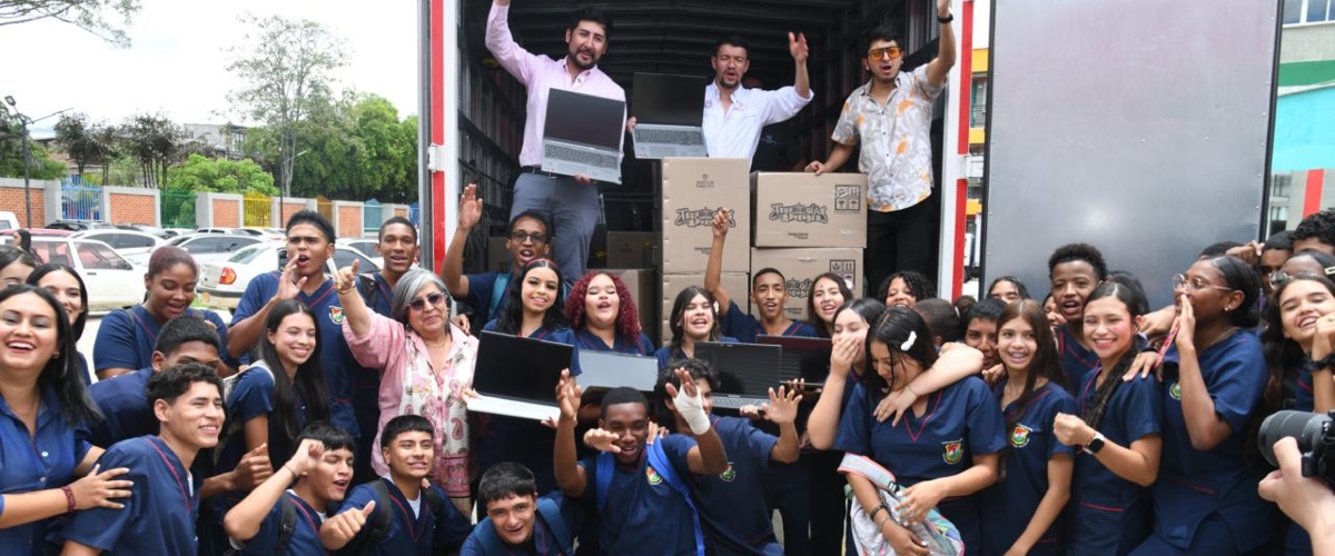 Grupo de estudiantes y adultos celebra frente a un camión abierto con cajas y portátiles, símbolo de entrega tecnológica para educación.