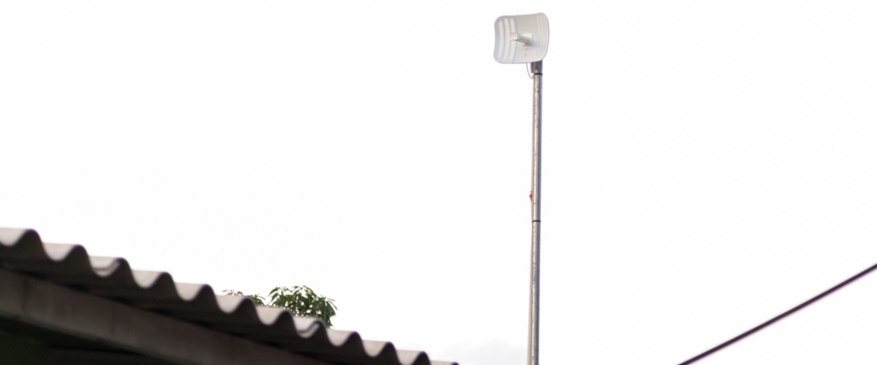 Antena de telecomunicaciones elevada sobre un poste junto a un techo, simboliza conectividad y expansión de redes en zonas apartadas.