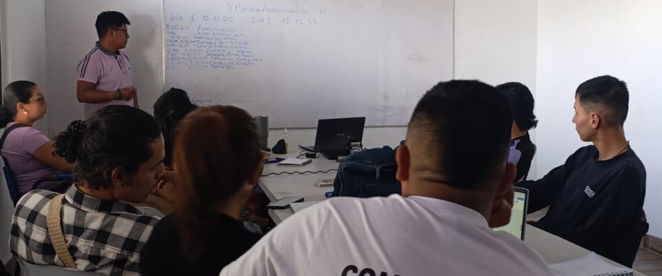 Grupo de personas en aula escucha a un expositor frente a tablero; escena educativa que evoca memoria y arraigo territorial