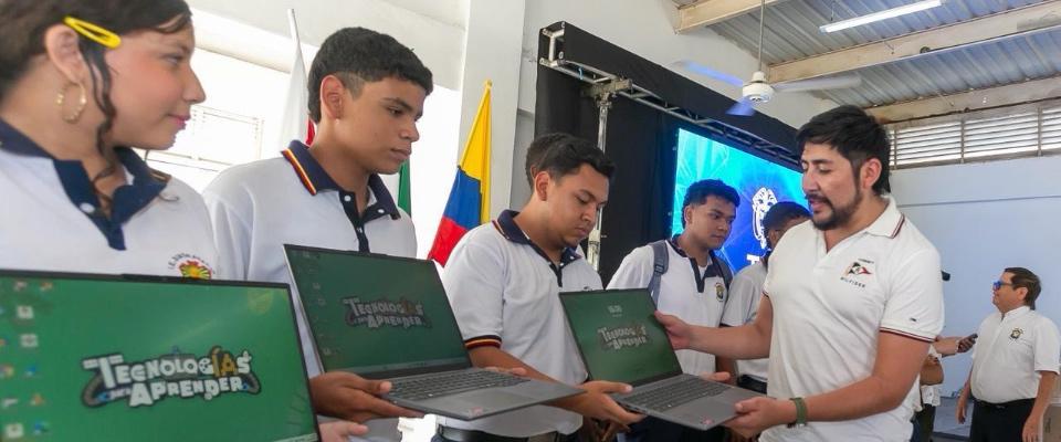 Estudiantes con uniforme reciben portátiles de un instructor; al fondo, bandera de Colombia y pantalla, impulso digital en Sucre