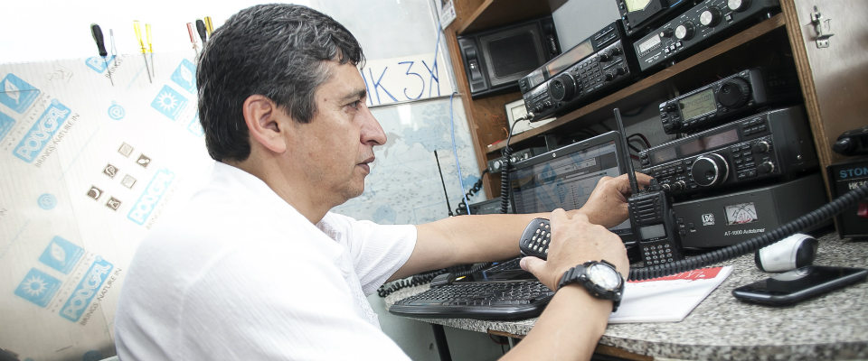 Hombre opera radios y equipos de comunicación en una mesa técnica, ajustando perillas y un walkie-talkie en entorno de radioafición.