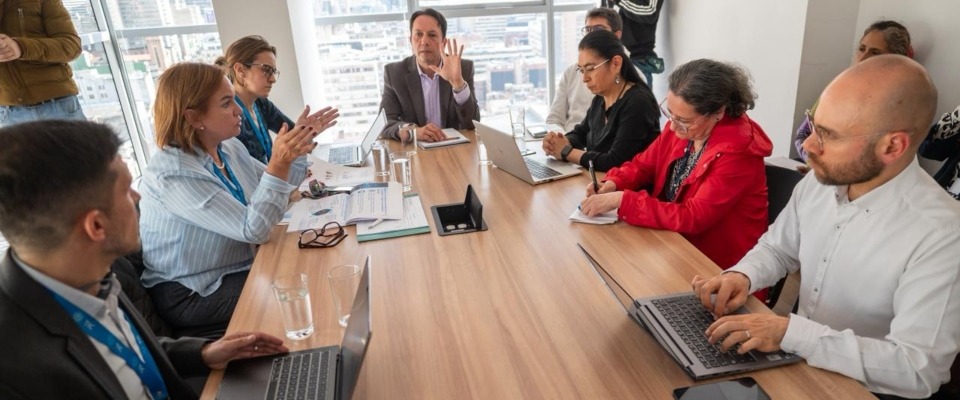 Grupo de profesionales en reunión discute temas técnicos; algunos usan portátiles y toman notas en mesa de juntas con vista urbana