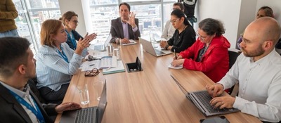 Grupo de profesionales en reunión discute temas técnicos; algunos usan portátiles y toman notas en mesa de juntas con vista urbana