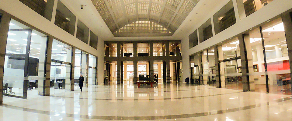 Amplio vestíbulo moderno con pisos brillantes, techos altos y locales a los lados. Pocas personas caminan en un espacio iluminado y ordenado.