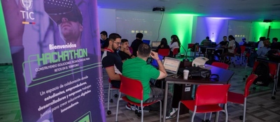 Personas trabajan en laptops durante hackathon TIC; mesas con equipos y luces verdes. Evento Colombia 5.0 reúne expertos y talleres