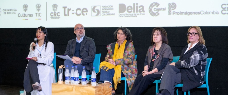 Cinco panelistas en escenario dialogan ante logos institucionales; una mujer habla con micrófono en foro audiovisual público.