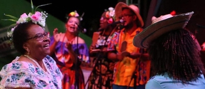 Mujer sonriente con flores en el cabello disfruta música en vivo; detrás, grupo canta y toca con vestuario colorido afro