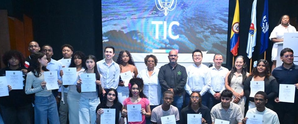 Grupo de jóvenes y adultos sostiene certificados en tarima; detrás, pantalla de MinTIC y banderas. Evento destaca liderazgo femenino.