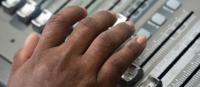 Primer plano de una mano operando una consola de audio. La imagen representa la radiodifusión comunitaria y la comunicación local.