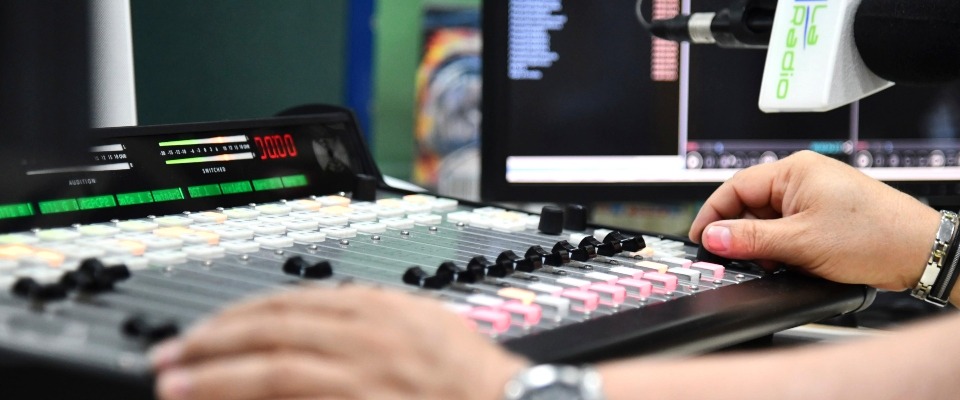 Persona opera una consola de audio en una cabina de radio, ajustando controles frente a monitores y un micrófono profesional.
