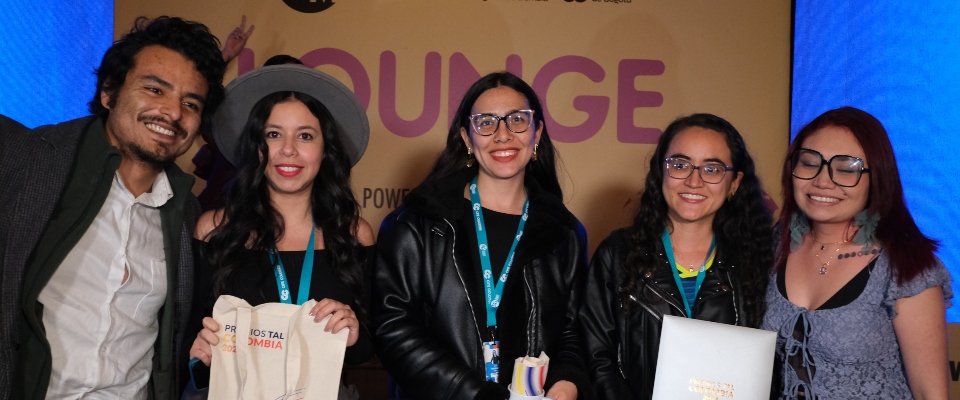 Cinco asistentes sonríen frente al panel de un evento, con acreditaciones y materiales, tras anuncio de nominados a Premios TAL. Cinco asistentes sonríen frente al panel de un evento, con acreditaciones y materiales, tras anuncio de nominados a Premios TAL.