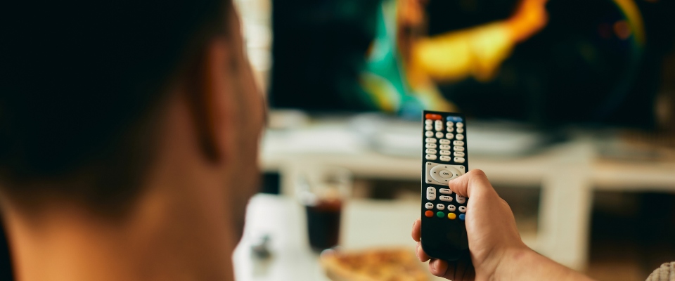 Persona de espaldas sostiene un control remoto frente a un televisor desenfocado. Al fondo hay un vaso y un plato sobre una mesa Persona de espaldas sostiene un control remoto frente a un televisor desenfocado. Al fondo hay un vaso y un plato sobre una mesa
