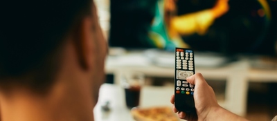 Persona de espaldas sostiene un control remoto frente a un televisor desenfocado. Al fondo hay un vaso y un plato sobre una mesa