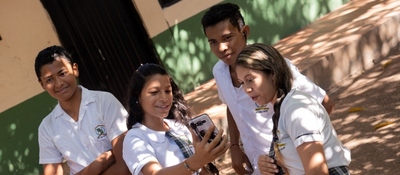 Cuatro estudiantes con uniforme blanco miran un celular en un patio con árboles; tres sonríen y uno observa de pie