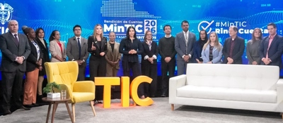 Catorce personas de pie detrás de sillón blanco, silla amarilla y letras TIC amarillas, frente a pantalla azul con textos