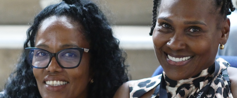 Dos personas sonríen a la cámara; izquierda con gafas negras, derecha con blusa estampada y arete dorado; fondo desenfocado. Dos personas sonríen a la cámara; izquierda con gafas negras, derecha con blusa estampada y arete dorado; fondo desenfocado.