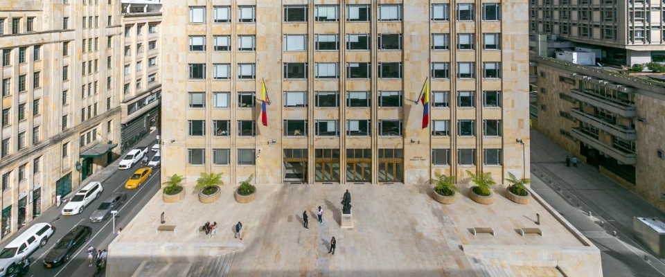 Edificio sede del Ministerio TIC con dos banderas, plaza de piedra, jardineras, estatua central, varias personas caminando y vehículos en calles laterales. Edificio sede del Ministerio TIC con dos banderas, plaza de piedra, jardineras, estatua central, varias personas caminando y vehículos en calles laterales.