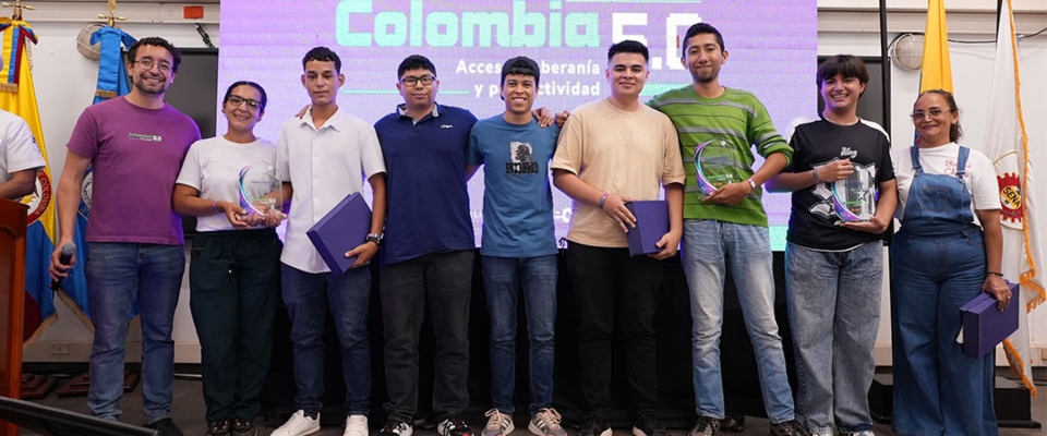 Nueve personas posan en tarima con placas y carpetas; al fondo, pantalla “Colombia 5.0” y banderas a ambos lados