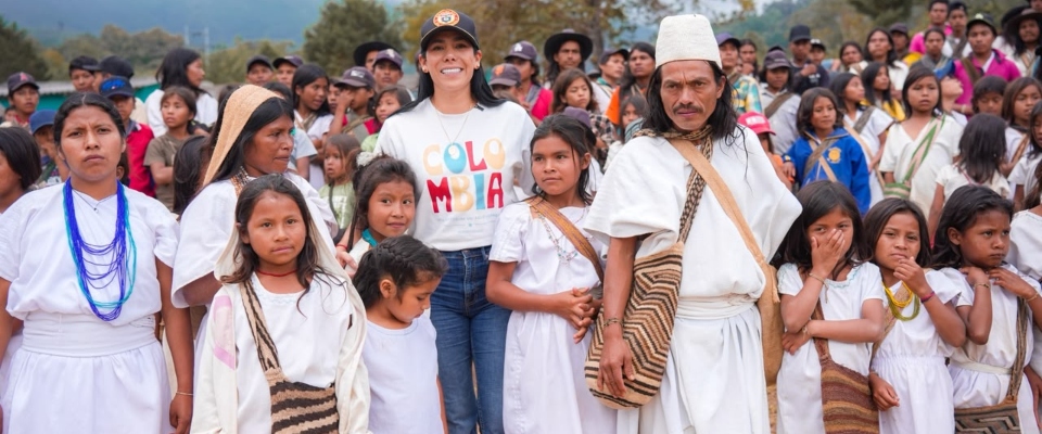 Grupo de personas con ropa blanca; mujer con camiseta "COLOMBIA" al centro. Niñas y niños alrededor, multitud al fondo.