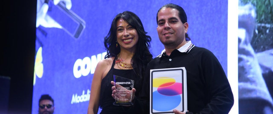 Dos personas sonríen en un escenario; una sostiene un trofeo y la otra una tableta. Fondo azul con pantalla y un asistente al fondo