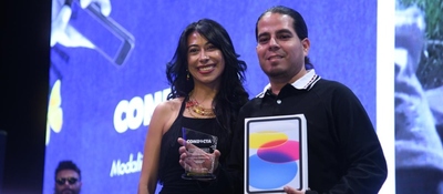 Dos personas sonríen en un escenario; una sostiene un trofeo y la otra una tableta. Fondo azul con pantalla y un asistente al fondo