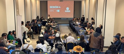 Sala con personas sentadas alrededor de mesas en U; al fondo, pantalla naranja con “Sesión 4 MPC 2026”; una persona expone.