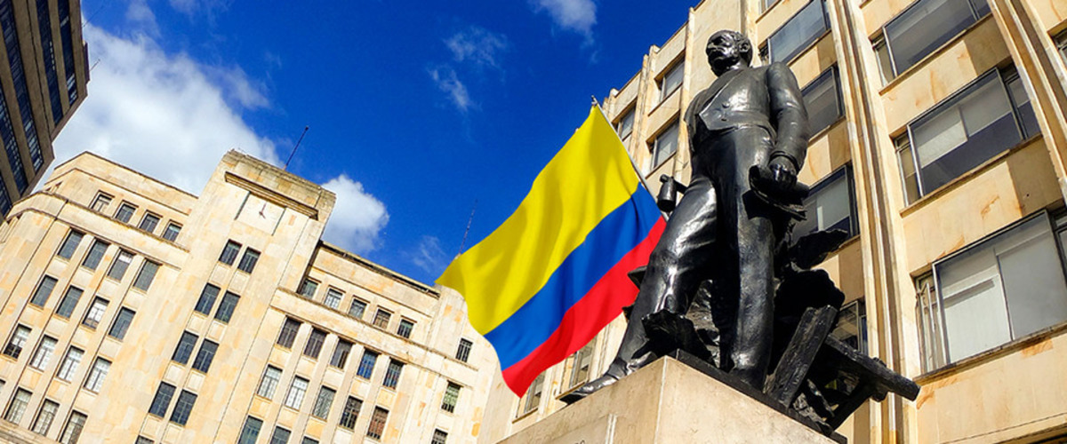 Estatua de bronce frente a edificios de piedra, con bandera de Colombia al centro y cielo azul con nubes. Estatua de bronce frente a edificios de piedra, con bandera de Colombia al centro y cielo azul con nubes.