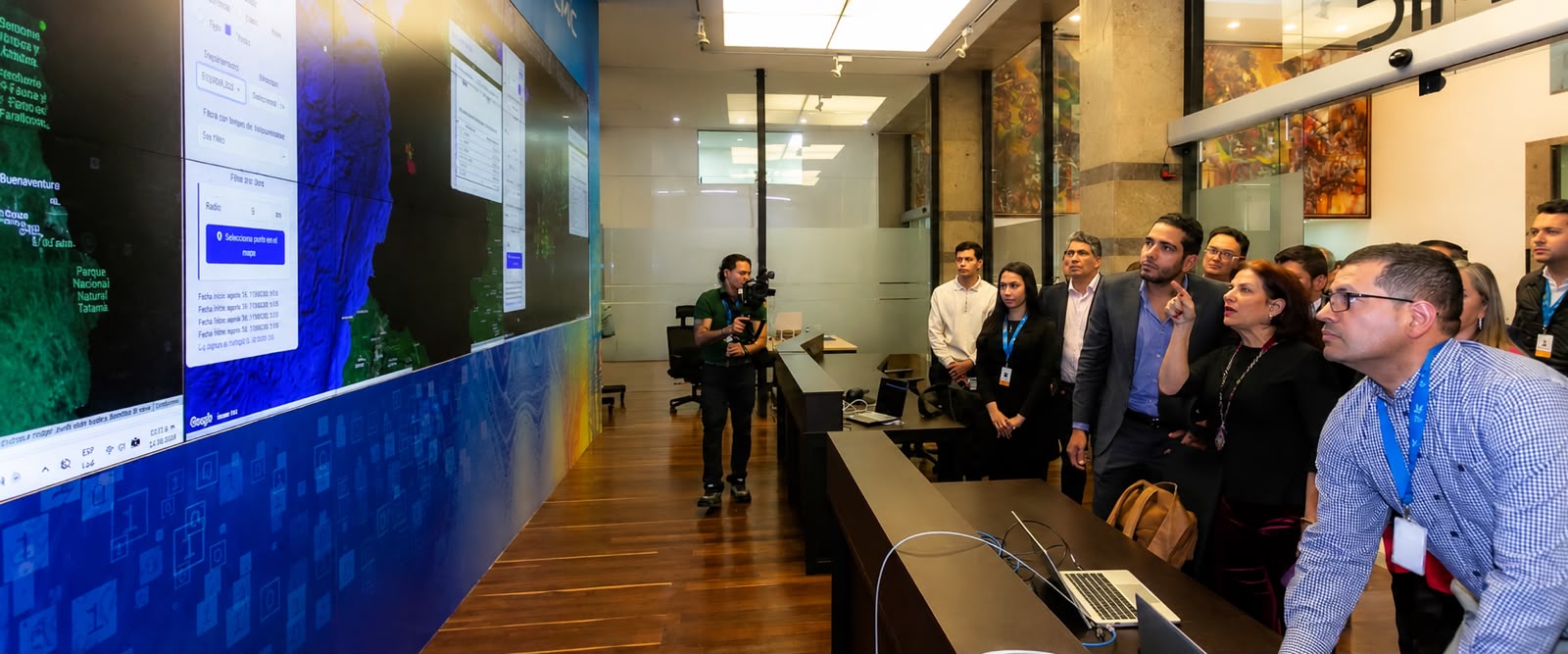 Personas observan pantallas con mapas y datos en una sala de monitoreo; hay mesas, computadores y cámaras