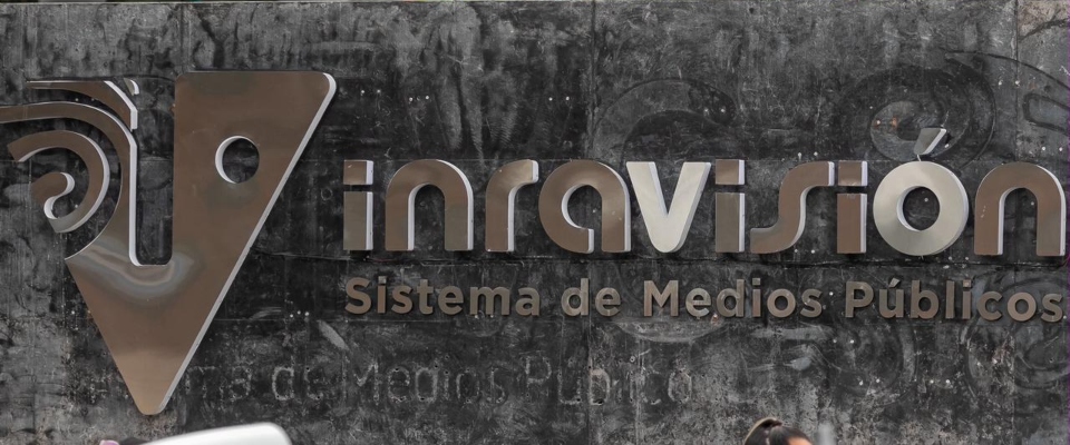 Logotipo de RTVC en tonos gris y beige sobre una pared oscura