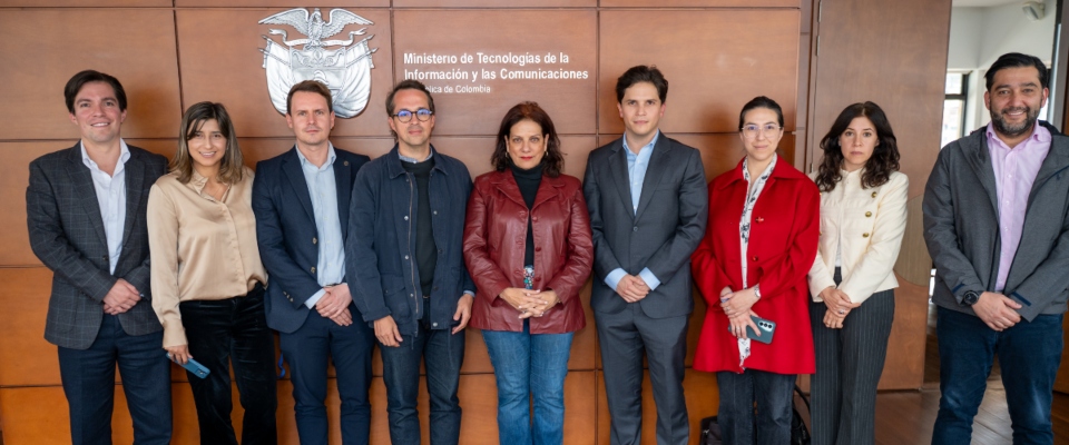 Nueve personas posan de pie frente al logo y nombre del Ministerio TIC; visten prendas en tonos oscuros, rojo, beige y gris Nueve personas posan de pie frente al logo y nombre del Ministerio TIC; visten prendas en tonos oscuros, rojo, beige y gris