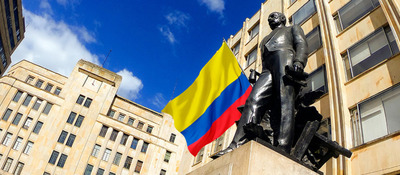 Estatua de una persona frente a edificios, con una bandera de Colombia ondeando al fondo bajo un cielo azul.