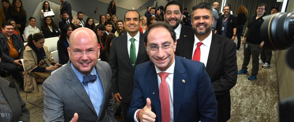 Cinco hombres con traje sonríen y dos hacen pulgar arriba; detrás hay asistentes sentados y de pie en una sala. Cinco hombres con traje sonríen y dos hacen pulgar arriba; detrás hay asistentes sentados y de pie en una sala.